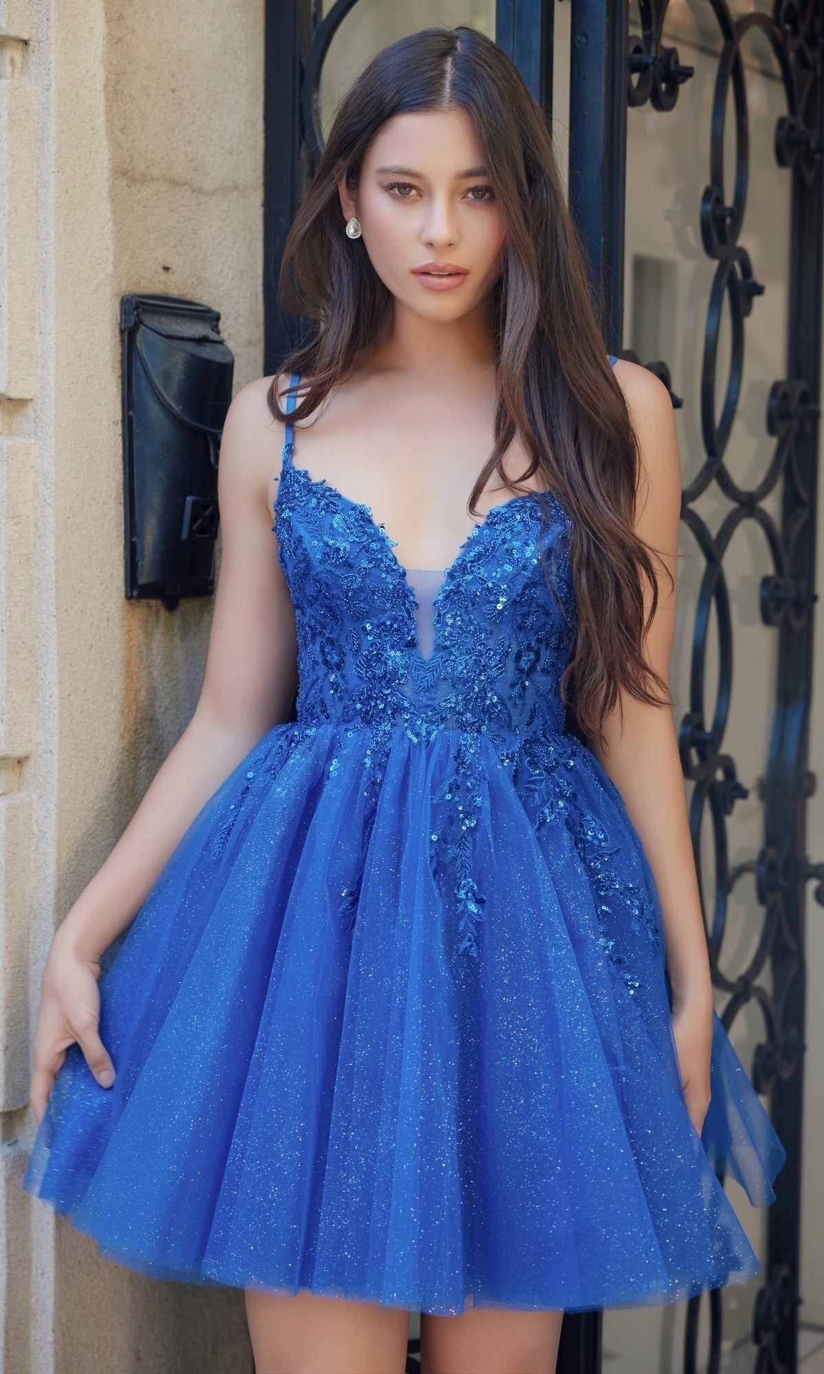 A woman stands before a wrought iron gate in the Nox Anabel E902 Glitter Tulle Homecoming Dress, a short blue design with sequin embroidery, floral embellishments, thin straps, and sparkling glitter tulle.